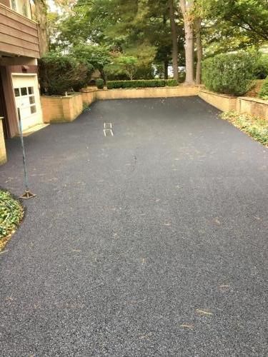 Asphalt driveway leading to a garage and a green backyard with trees and shrubs.