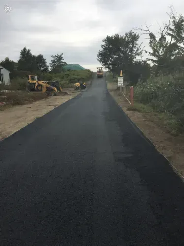Newly paved asphalt road; construction vehicles visible at a distance. Cloudy sky, trees, and roadside vegetation.