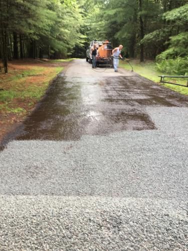 Asphalt being sprayed on a gravel driveway. Two workers near a tank, one spraying. Forest background.