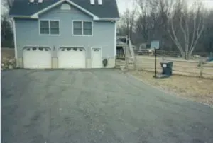 Two-story blue house with three-car garage. Asphalt driveway slopes up to the house. Basketball hoop in the yard.