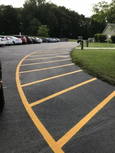 Yellow painted lines on a paved curve approaching a parking lot with cars.