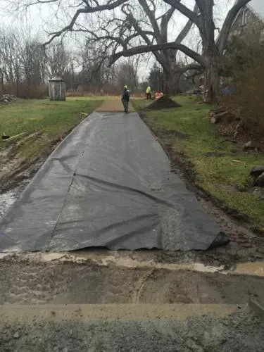 Construction site: Long black fabric laid on a driveway, worker walking on it, equipment and workers in the background.