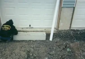 Person kneeling, working on a concrete structure near a garage door. Gravel and foundation visible.