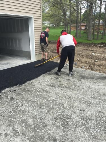 Two people paving a driveway with asphalt next to a garage.
