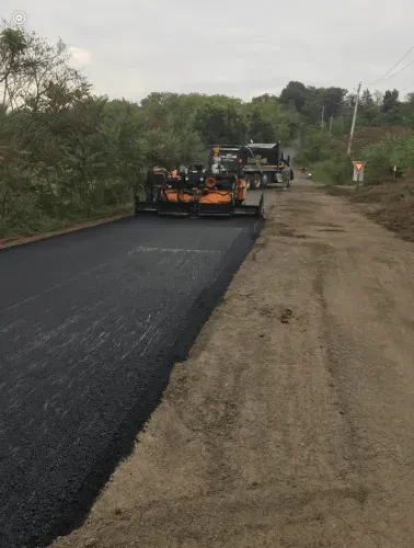 Asphalt paving machine laying blacktop on a dirt road, trees in the background.