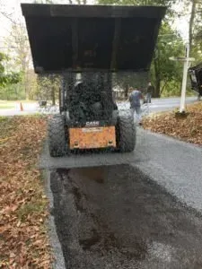 Skid steer with raised bucket spreading fresh asphalt on a driveway. A person is visible in the background.
