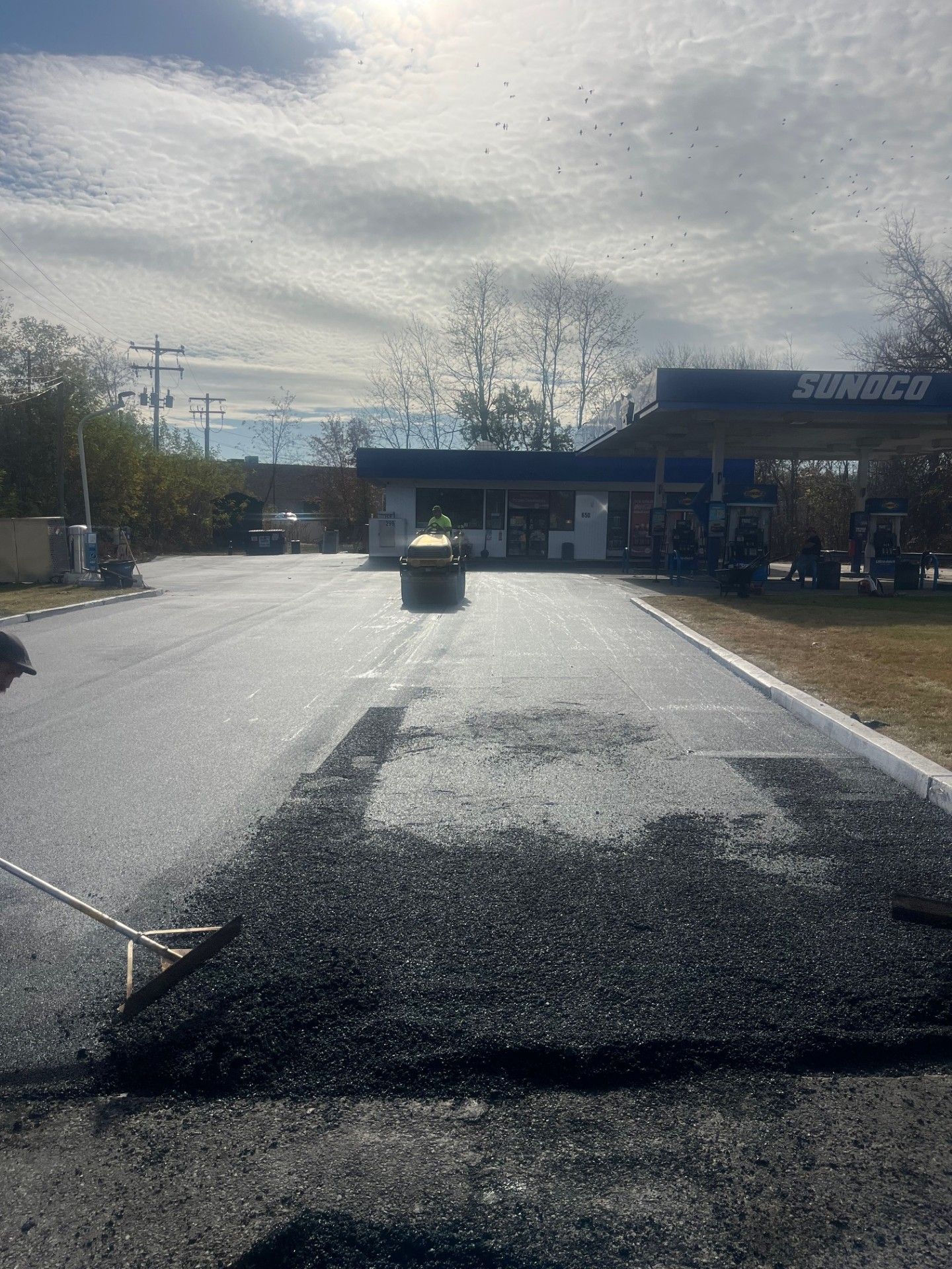 Asphalt paving at a Sunoco gas station. Black asphalt, gravel, a yellow barrel, and a cloudy sky.