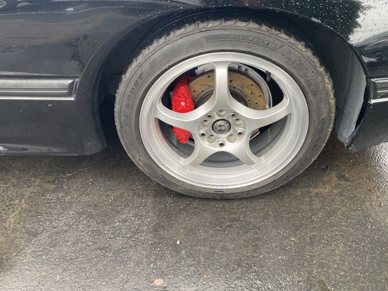 Close-up of a black car’s rear wheel with silver rim and red brake caliper on wet pavement