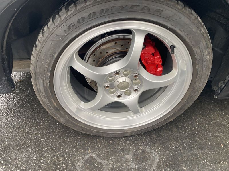 Silver car wheel with red brake caliper and drilled brake rotor on pavement