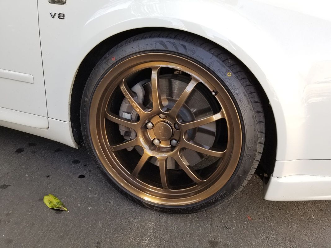 Bronze alloy wheel on a white car, parked on pavement.