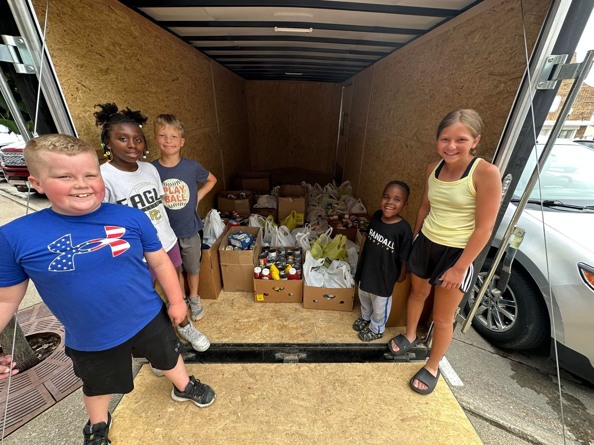 A group of children are standing in front of a trailer filled with boxes of food.