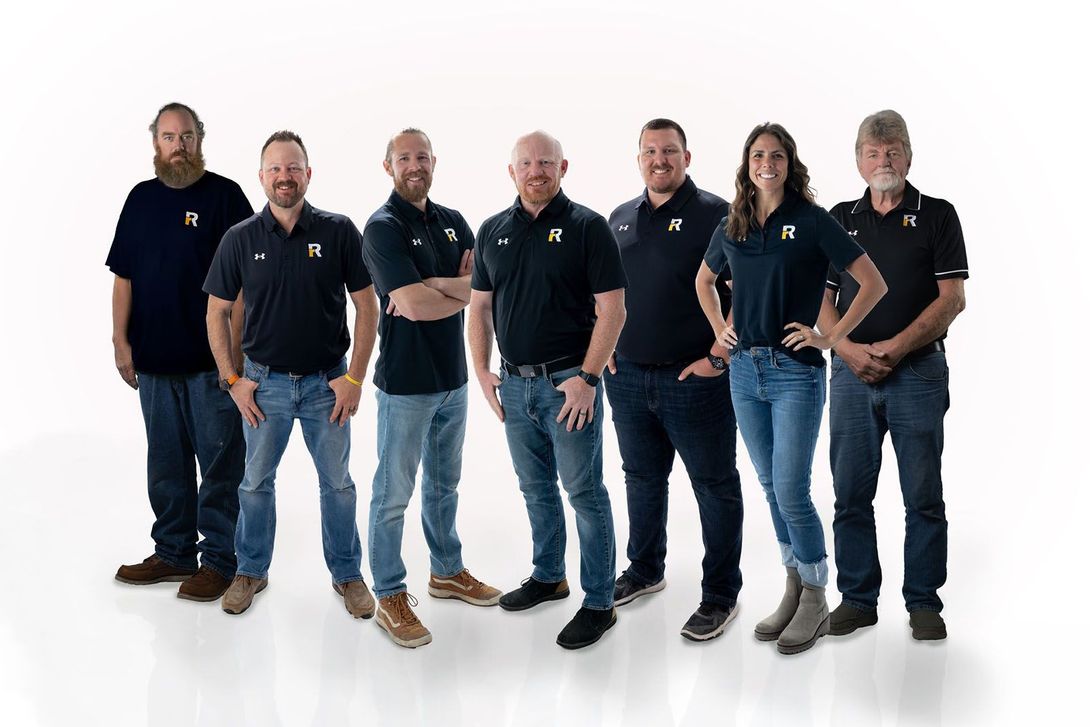 Group of eight people, wearing black shirts and jeans, posing against a white background.