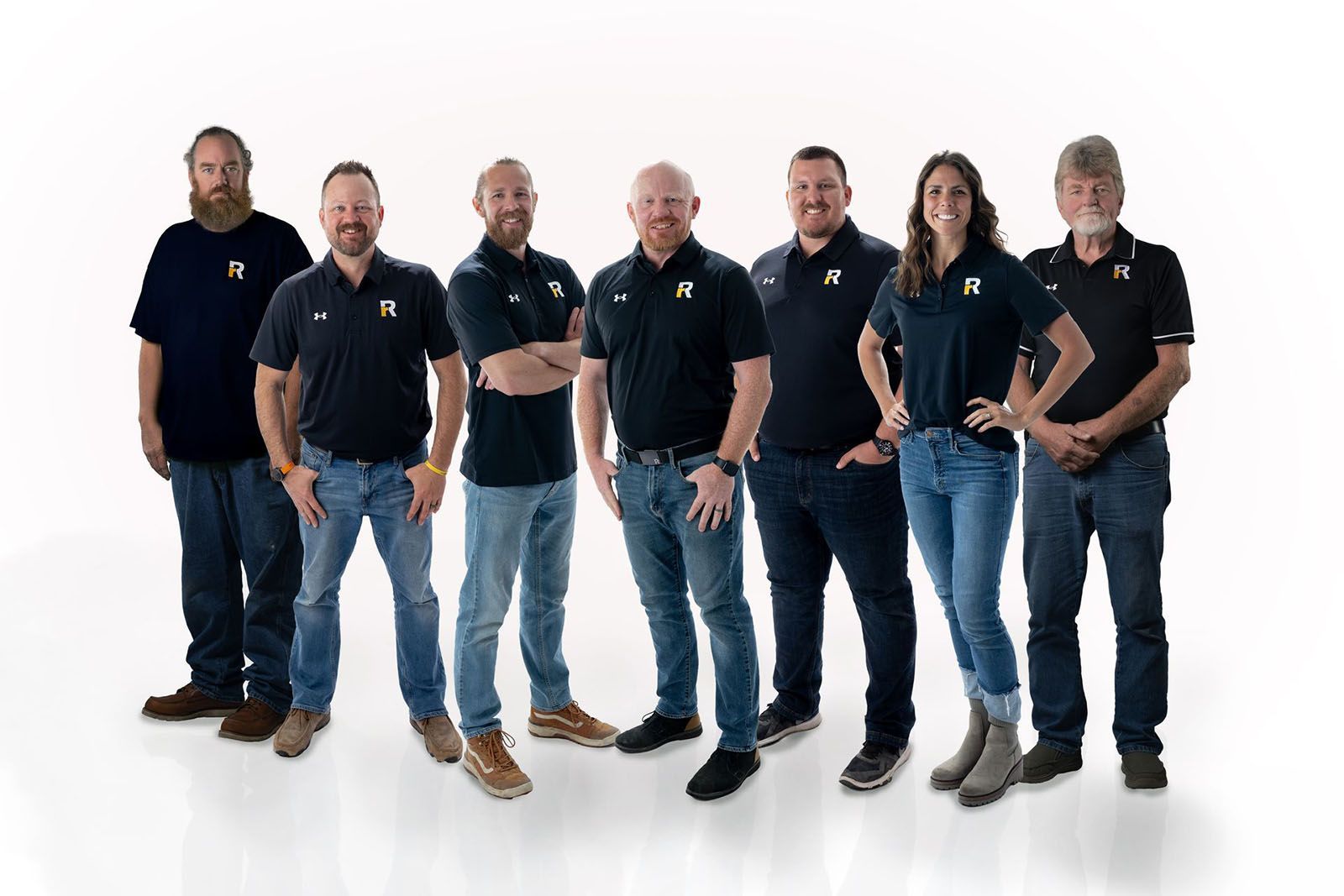 Group of eight people, wearing black shirts and jeans, posing against a white background.