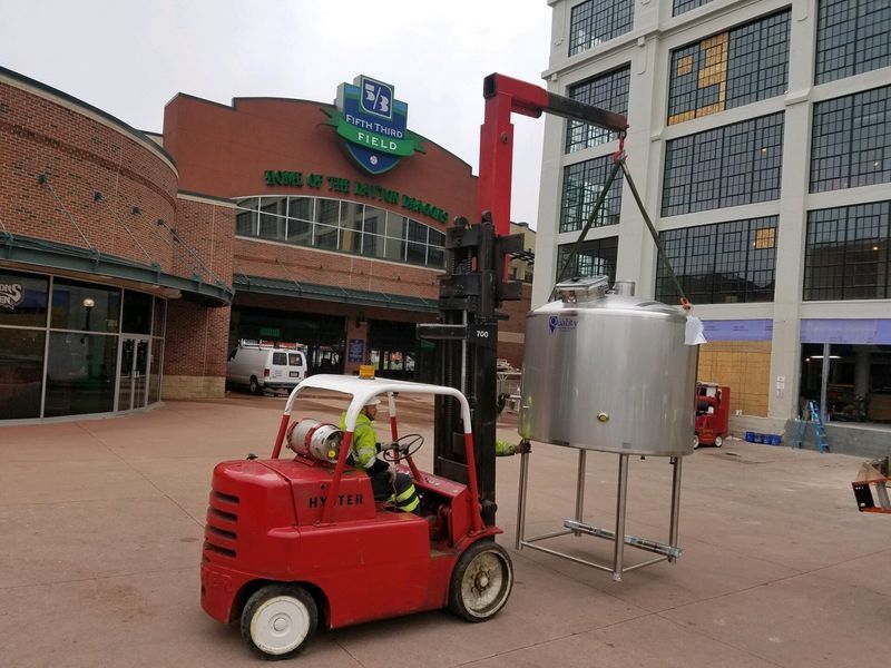 Red forklift lifting a large silver tank outside a brick building with a sign that reads 
