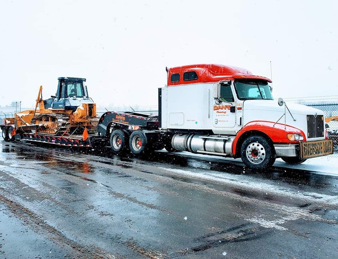 Truck hauling a yellow bulldozer on a snowy road. The truck has a white cab with red accents.