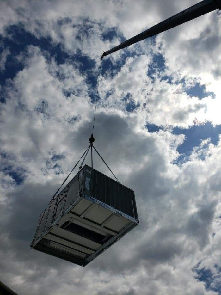 HVAC unit suspended in the air by crane cables against a cloudy sky.