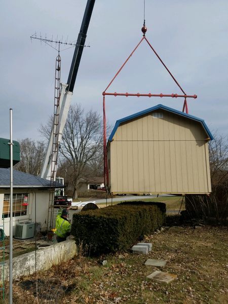 Crane lifting a tan shed with a blue roof, near a house and trees. A worker watches.