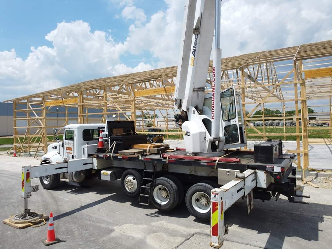 Truck-mounted crane at a construction site, lifting materials. Wooden framework of building visible. Bright sunny day.