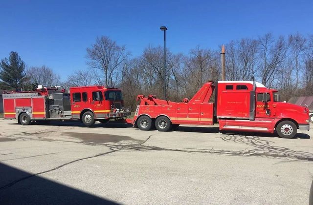 Red tow truck towing a red fire truck in a parking lot on a sunny day.