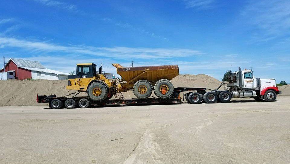 Truck hauling construction equipment on a flatbed trailer on a gravel lot under a blue sky.