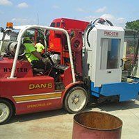 Red forklift carrying a white and blue machine outside; a person in a safety vest is operating it.