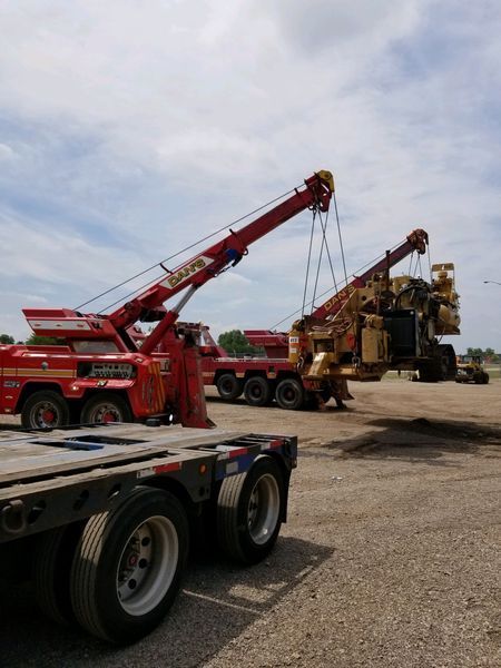 Two red tow trucks lifting a large, yellow industrial machine onto a flatbed trailer outdoors on a sunny day.