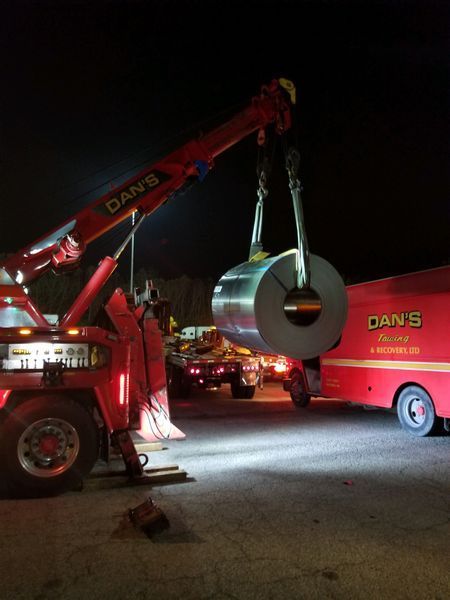 A red tow truck with a crane lifting a large metal coil at night. The truck has 