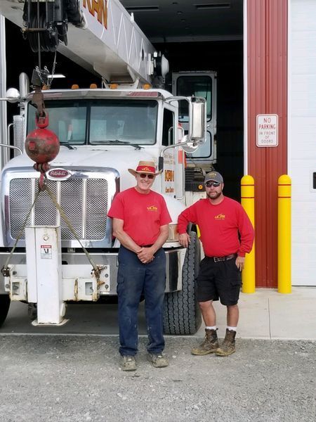 Two men in red shirts pose in front of a white crane truck in front of a red building with a loading dock.