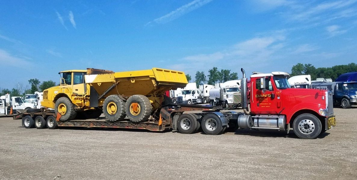 Red semi-truck hauling a large yellow dump truck on a flatbed trailer, under a blue sky.