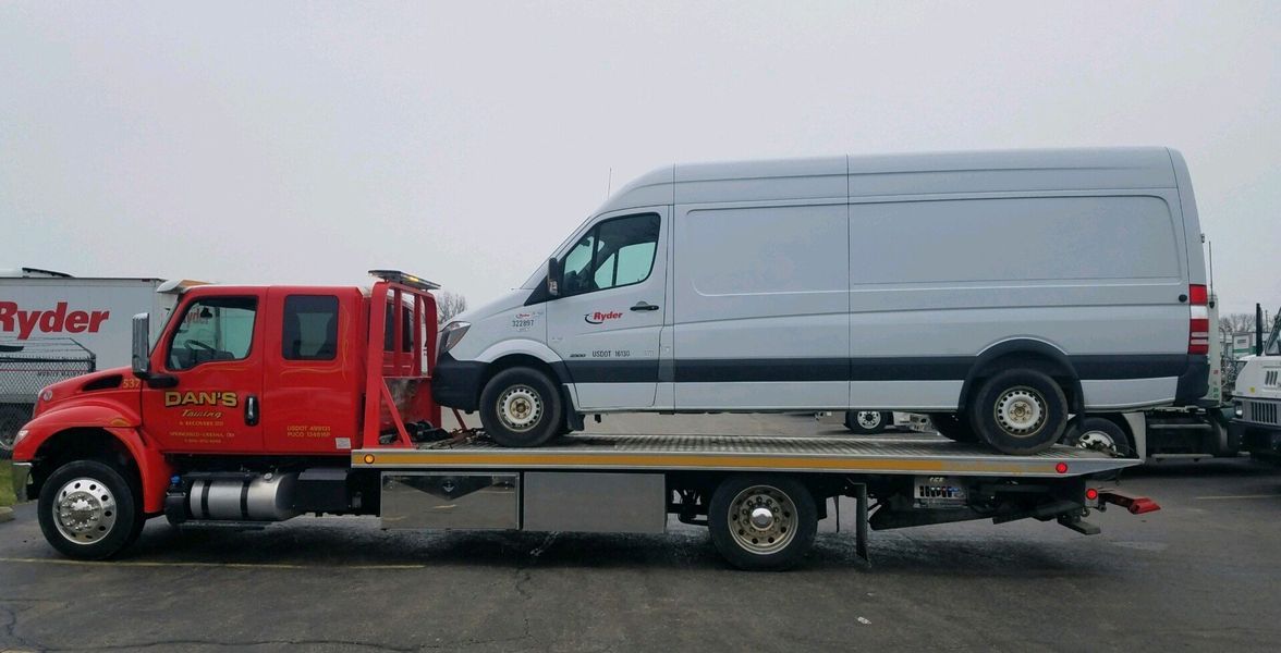 Red tow truck carrying a white cargo van on a flatbed trailer, parked outdoors.