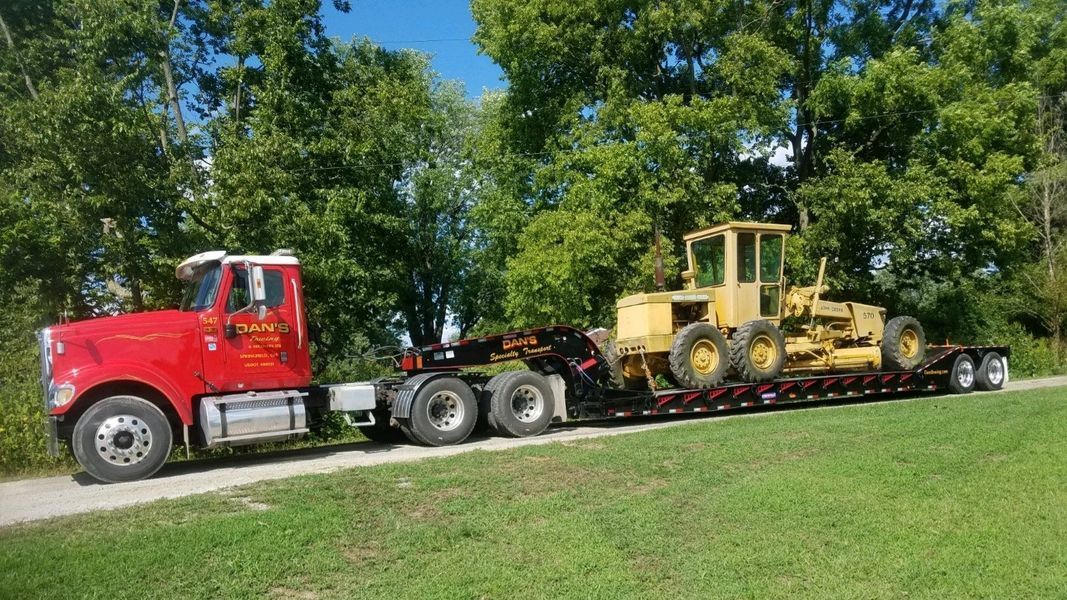 Red semi-truck hauling yellow road grader on a trailer, on a grassy road with trees in the background.