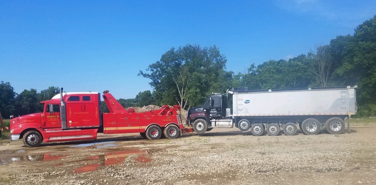 Red tow truck towing a dump truck on gravel with a forest in the background on a sunny day.