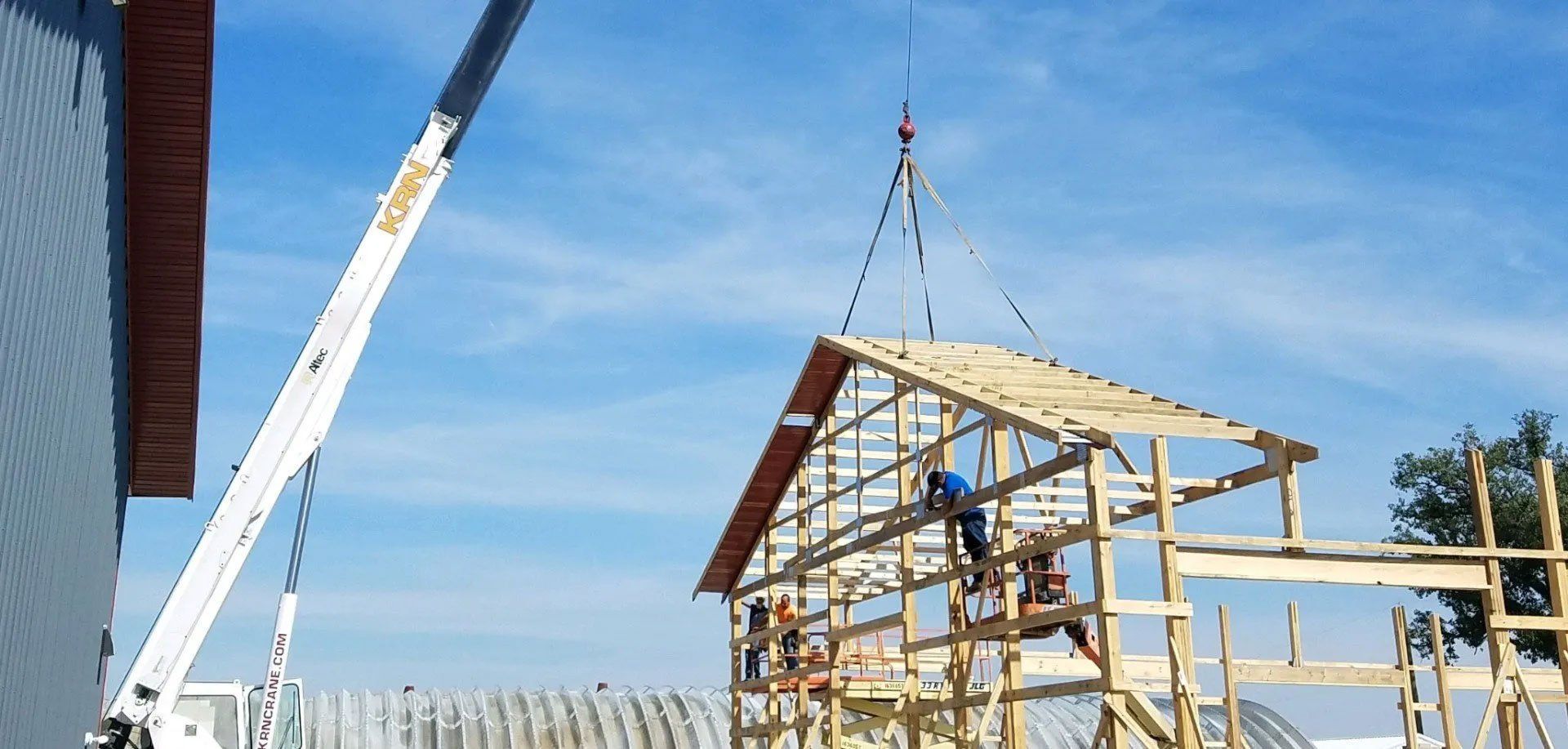 A crane lifts a barn roof into place as construction workers assemble the wooden frame under a blue sky.