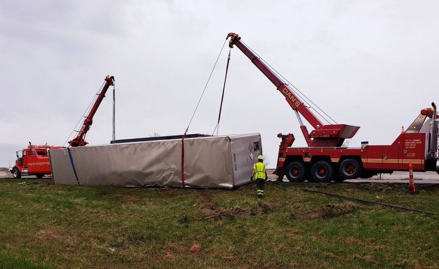 Two red tow trucks lifting a large, wrapped object on a grassy area, likely a roadside. A person in yellow stands nearby.