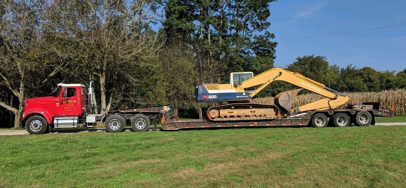 Red semi-truck hauling a yellow excavator on a trailer along a green grassy area.