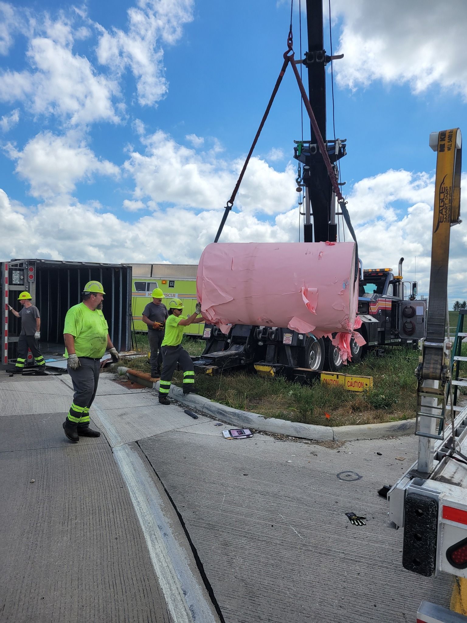 A crane lifting a damaged pink tank from a truck on the side of a road. Workers in safety gear. Bright blue sky.