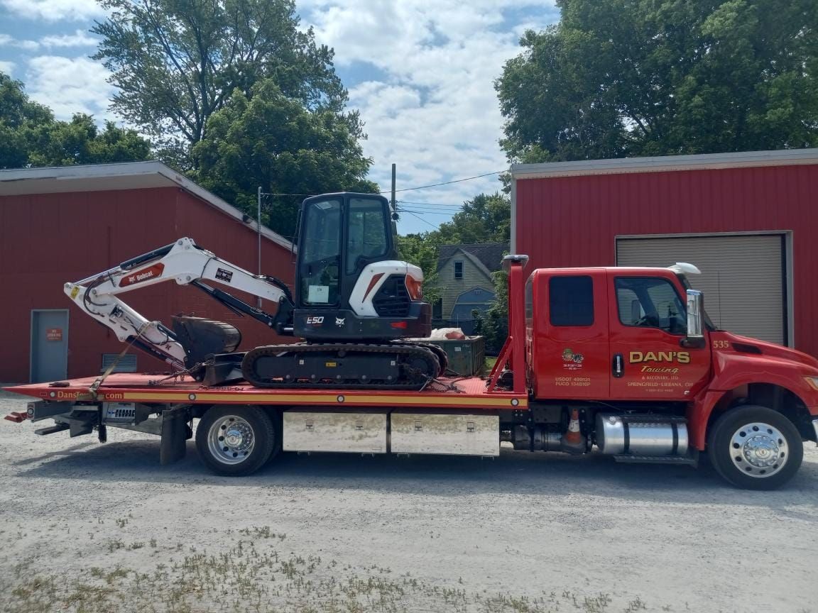 Red truck with a Bobcat excavator on its flatbed, parked in front of red buildings.