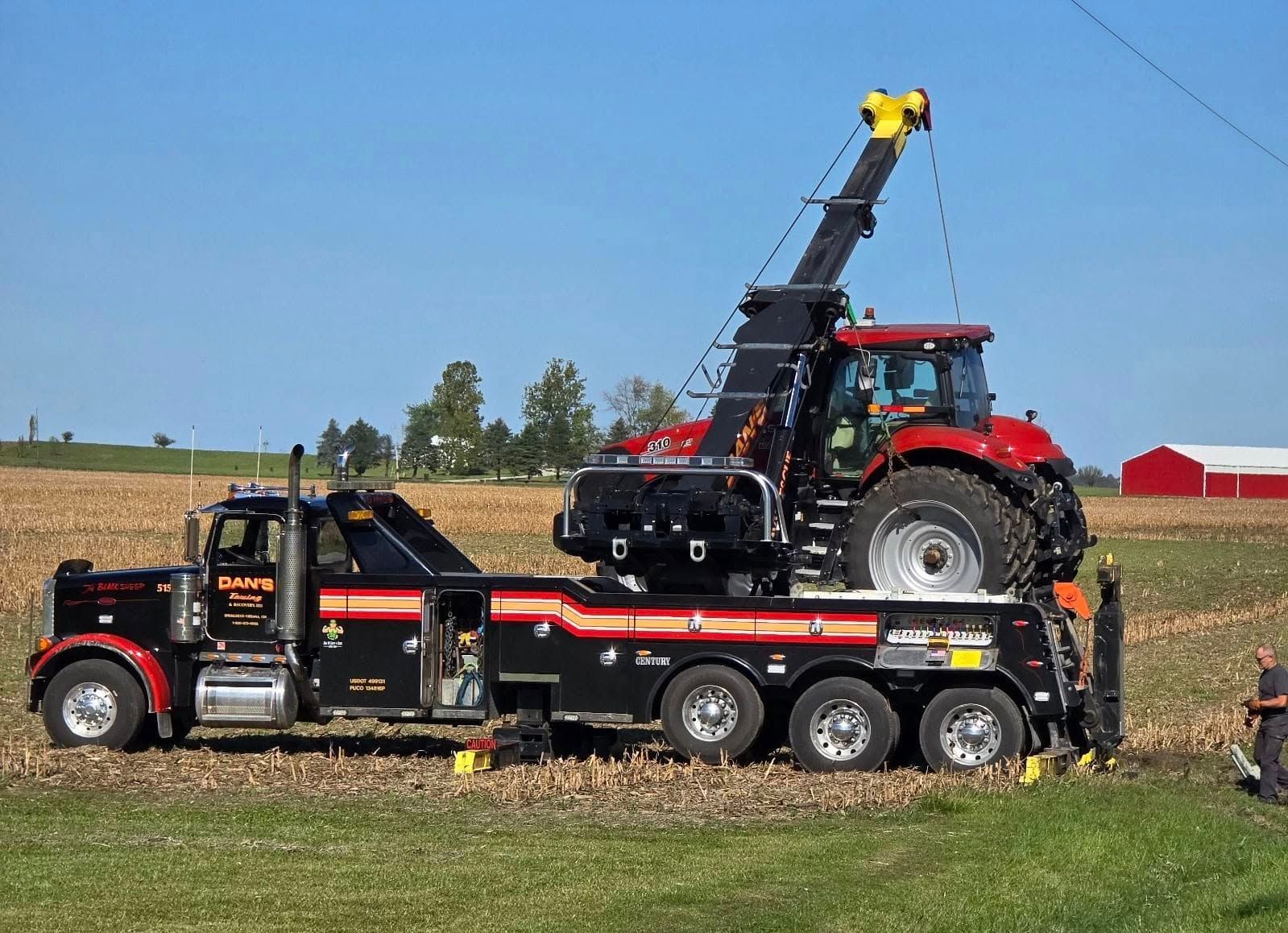 Tow truck lifting a red tractor from a field; black truck with extended crane.