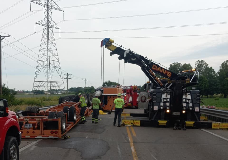 Tow truck lifting an overturned orange trailer on a road. Workers in yellow vests nearby; power lines in background.