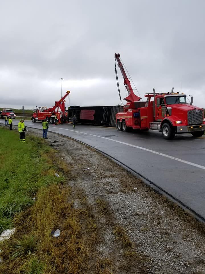 Two tow trucks righting a tipped semi-truck on a highway, roadside workers in safety vests.