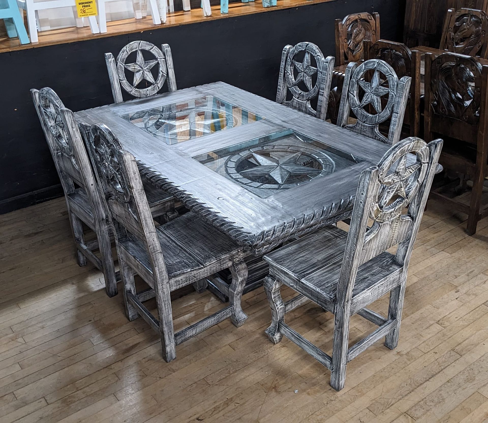 Rustic silver dining set with star accents. Table has glass inlay. Eight chairs surround it on a wooden floor.