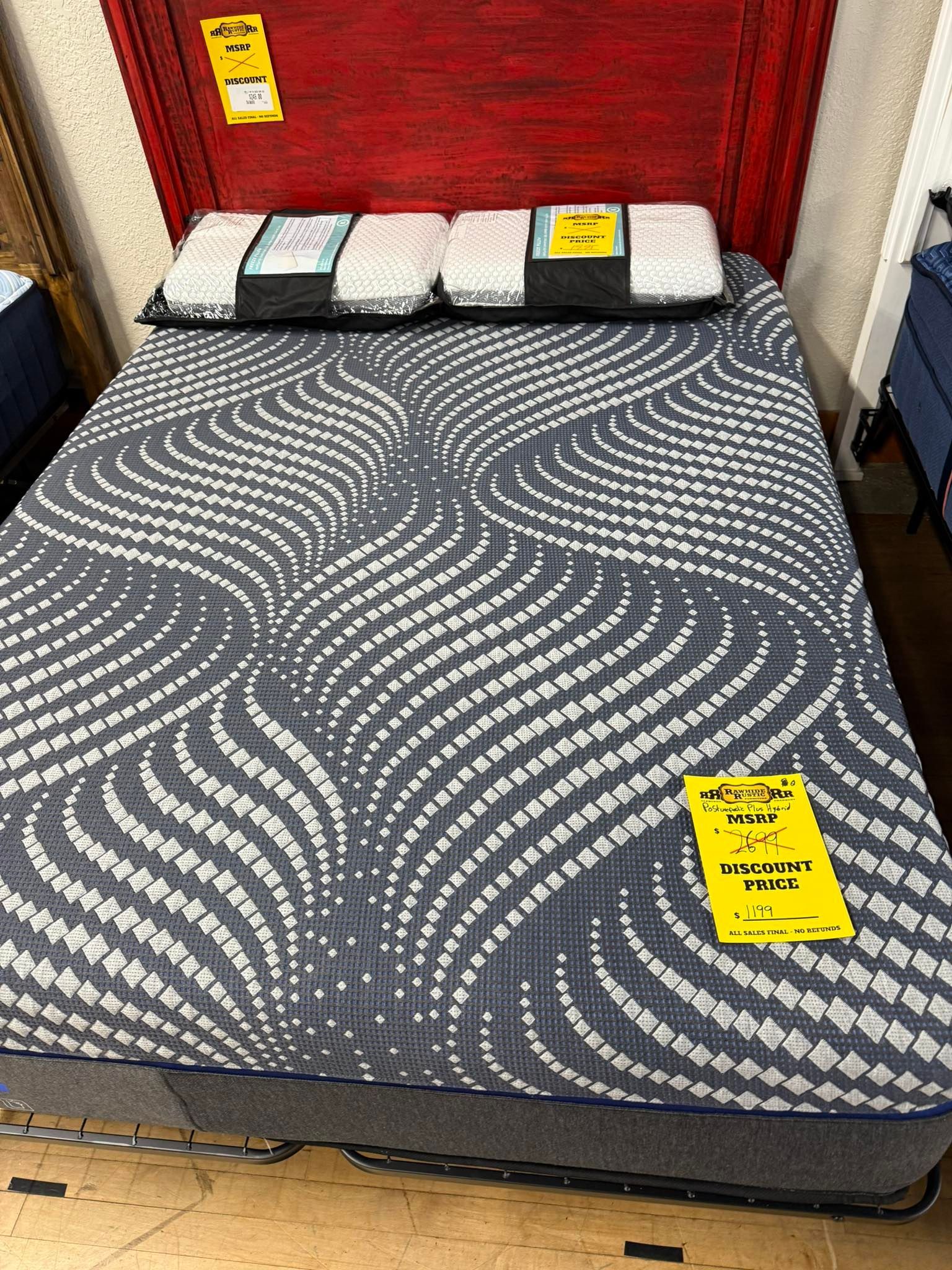 A display bed with gray patterned mattress, red headboard, and price tags.