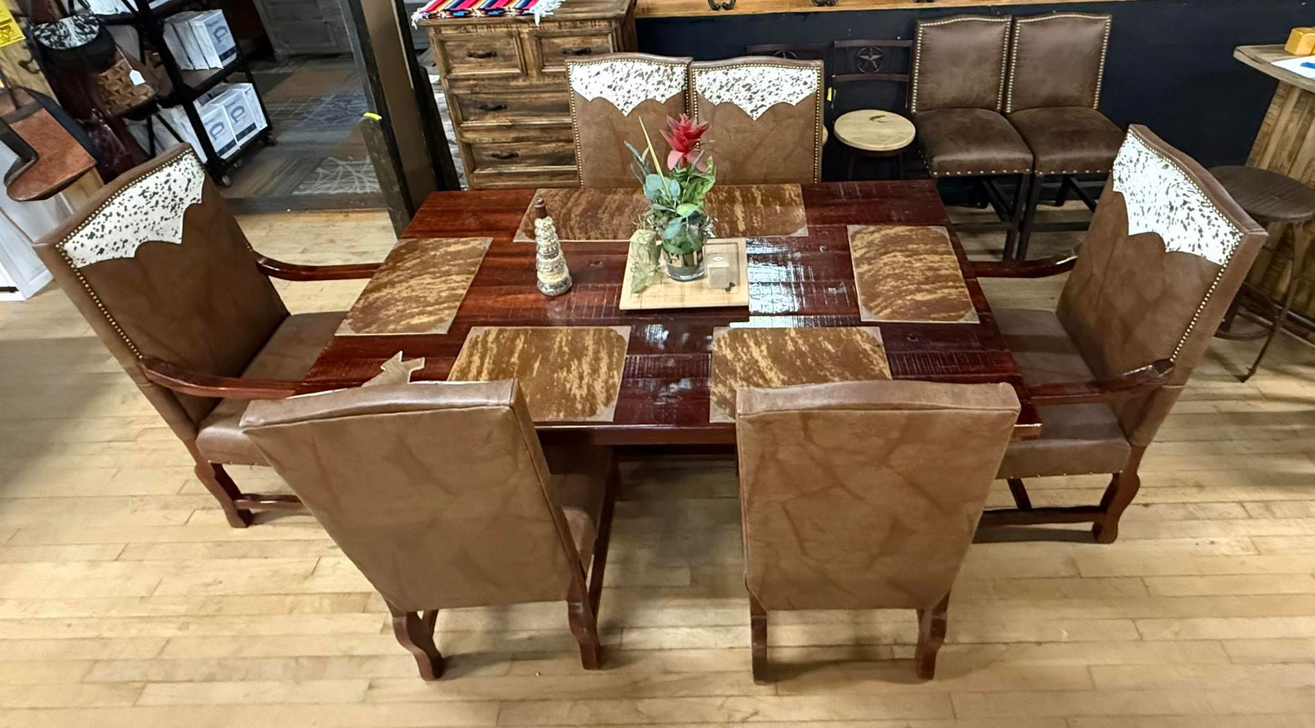 A rustic dining table with six leather chairs. The table has a wooden top with placemats and a centerpiece.
