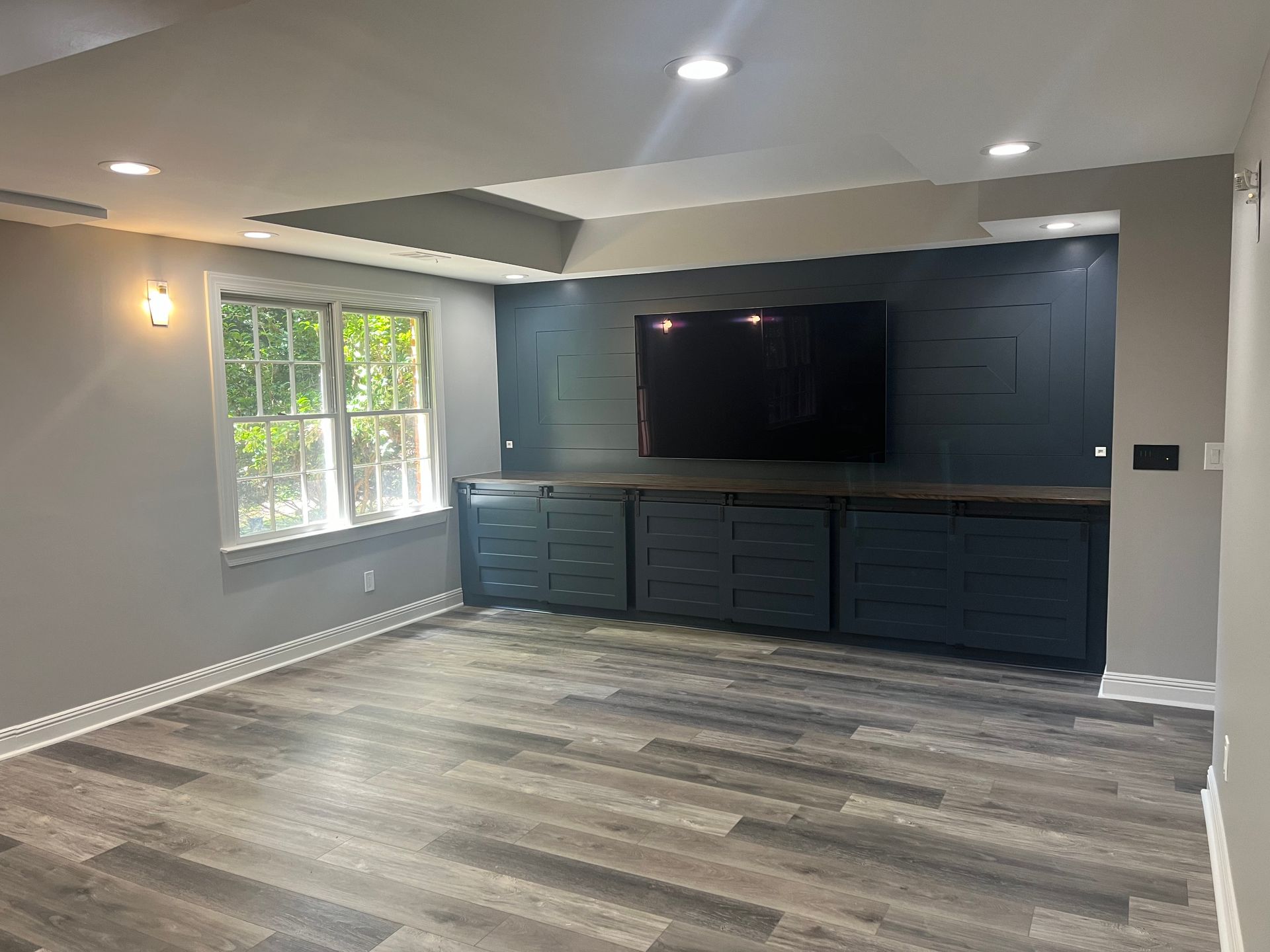 Empty room with blue built-in cabinets and TV, wood-look floor, gray walls, and window.