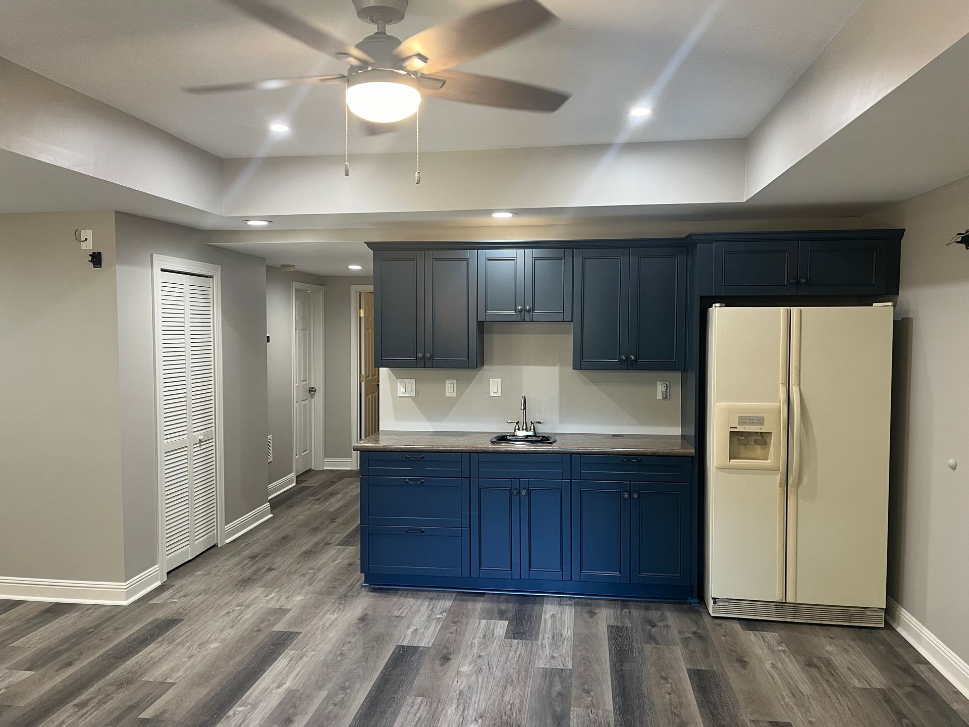 Basement kitchen with blue cabinets, a cream refrigerator, and gray wood-look flooring.