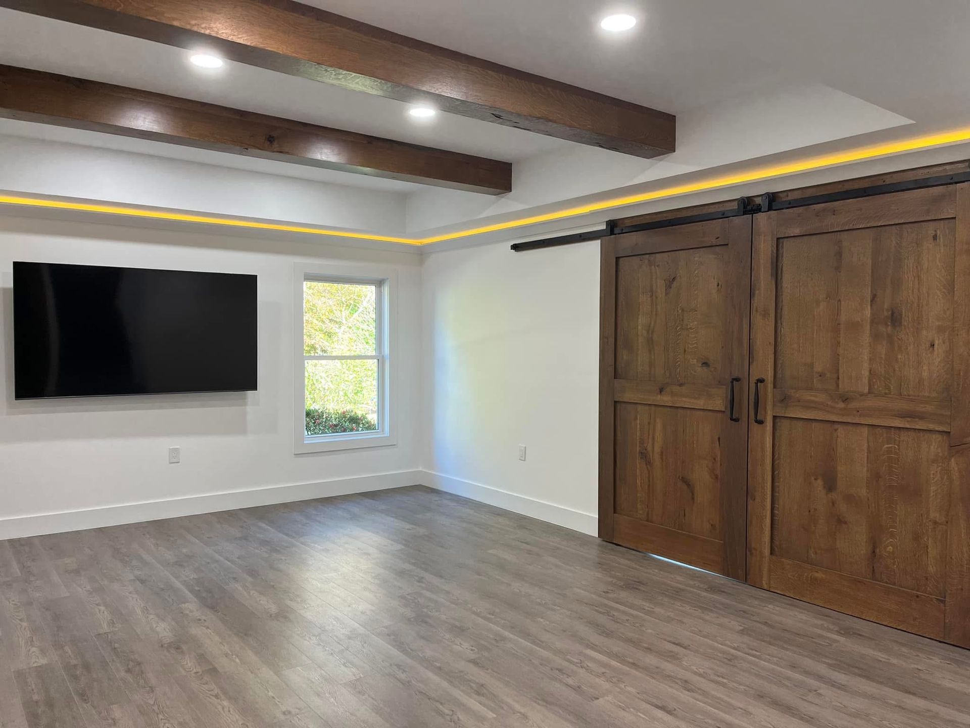 Empty room with wood-look sliding doors, beams, and flooring. A mounted TV is on the wall, next to a window.