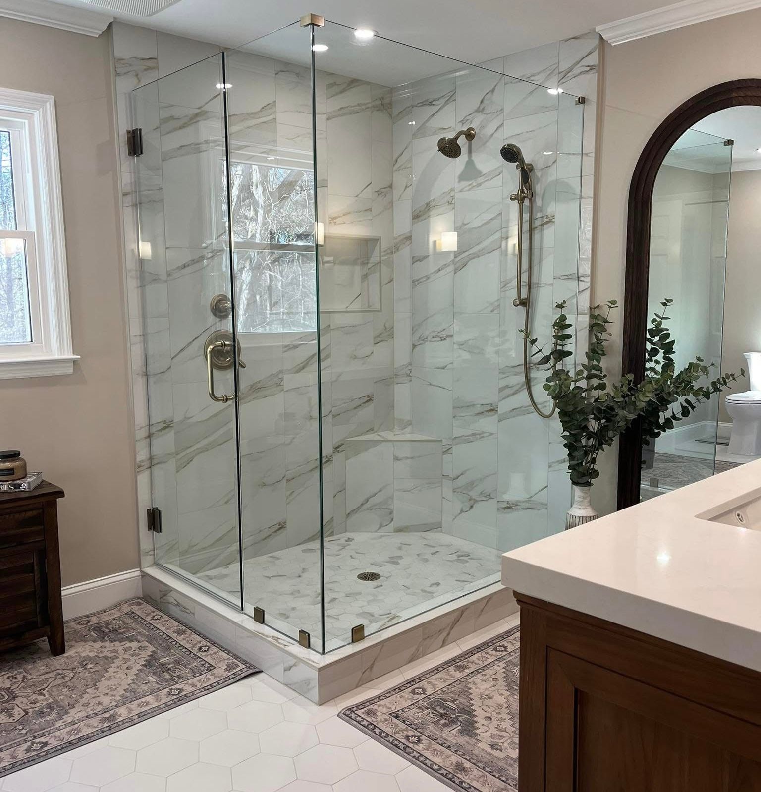 Glass-enclosed shower with marble-look tiles, brass fixtures, and a white base in a remodeled bathroom.