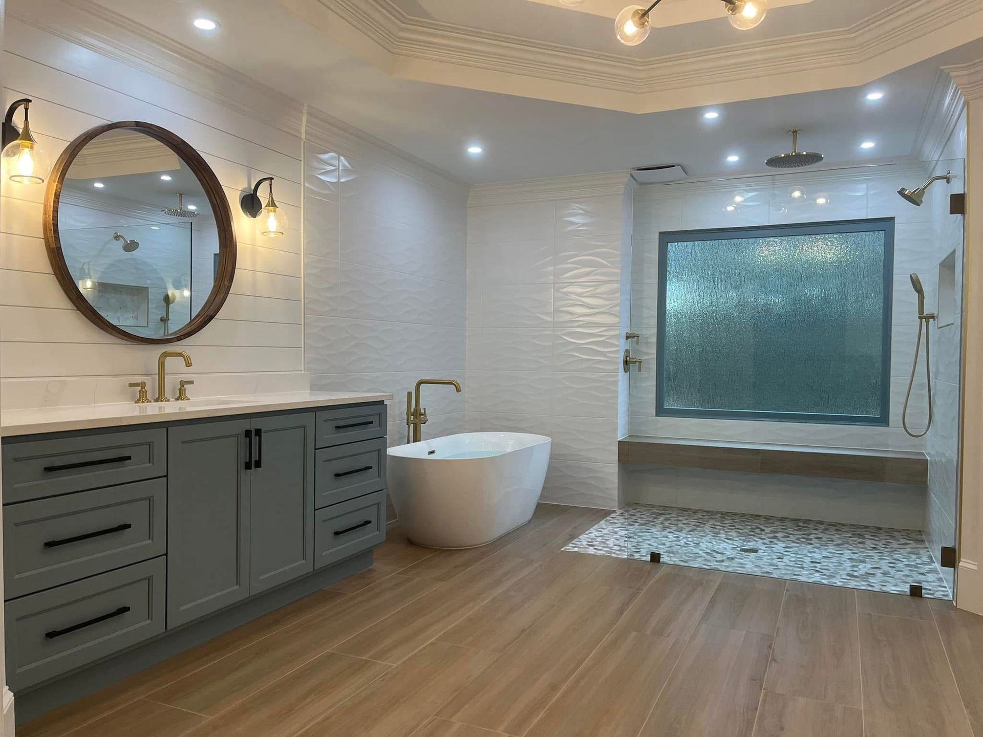 Luxurious bathroom with gray vanity, round mirror, white tub, and large glass shower.