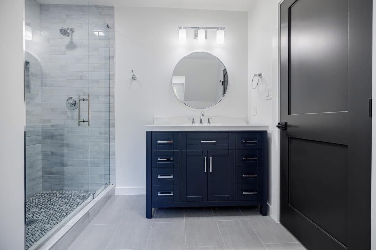 Bathroom with dark blue vanity, round mirror, glass shower with pebble floor, gray tile.