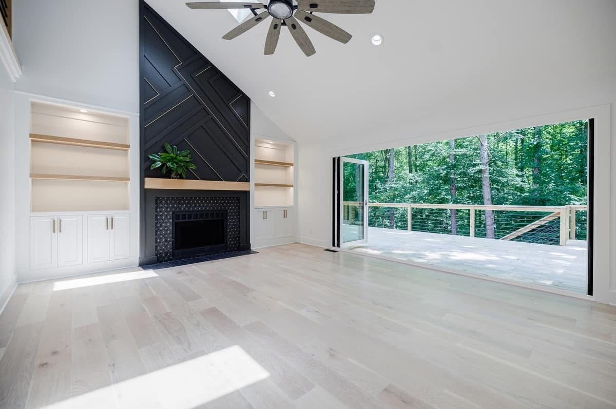 Bright living room with dark fireplace and open doors to a deck overlooking a wooded area.